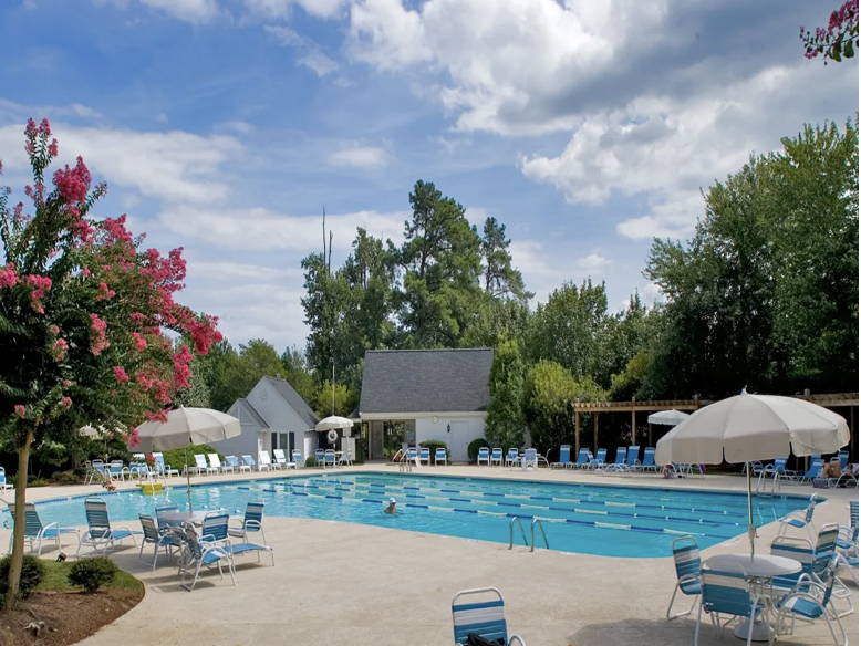 A serene outdoor swimming pool surrounded by lounge chairs and umbrellas. The pool is divided into lanes, with a few people swimming. In the background, there are trees, a small building, and a wooden pergola. The sky is partly cloudy. Fearrington Village